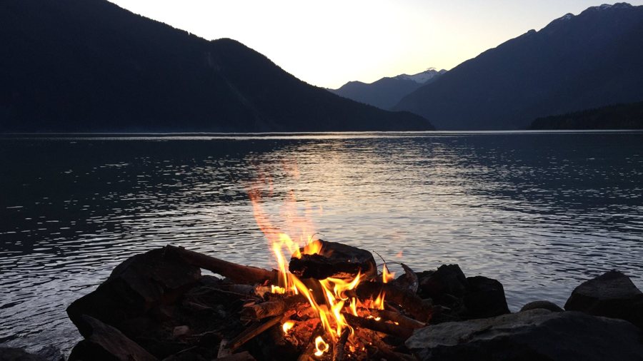 A campfire on a rocky shore, a lake and mountains behind at dusk